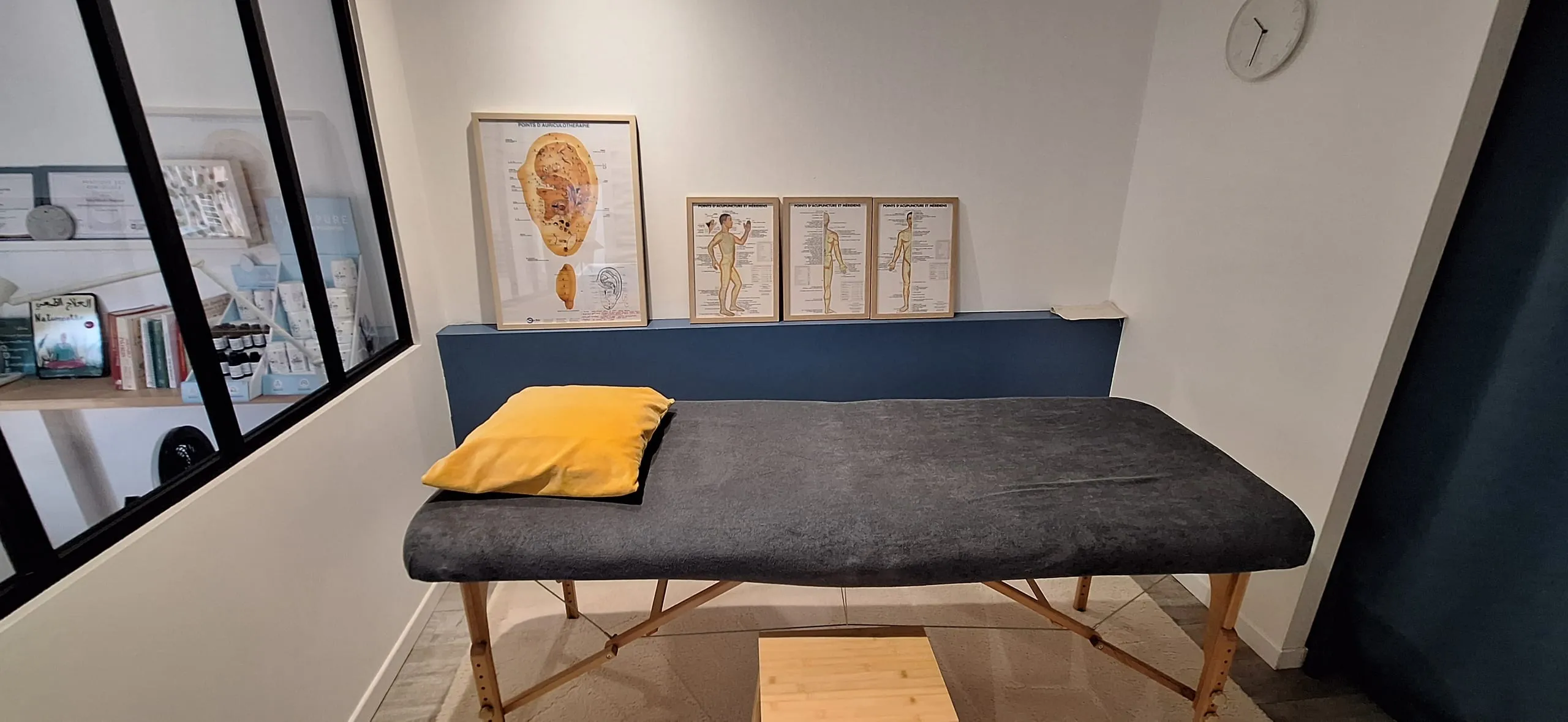 Salle de soin lumineuse avec table de massage, drap gris, coussin jaune et planches d’auriculothérapie au mur bleu.