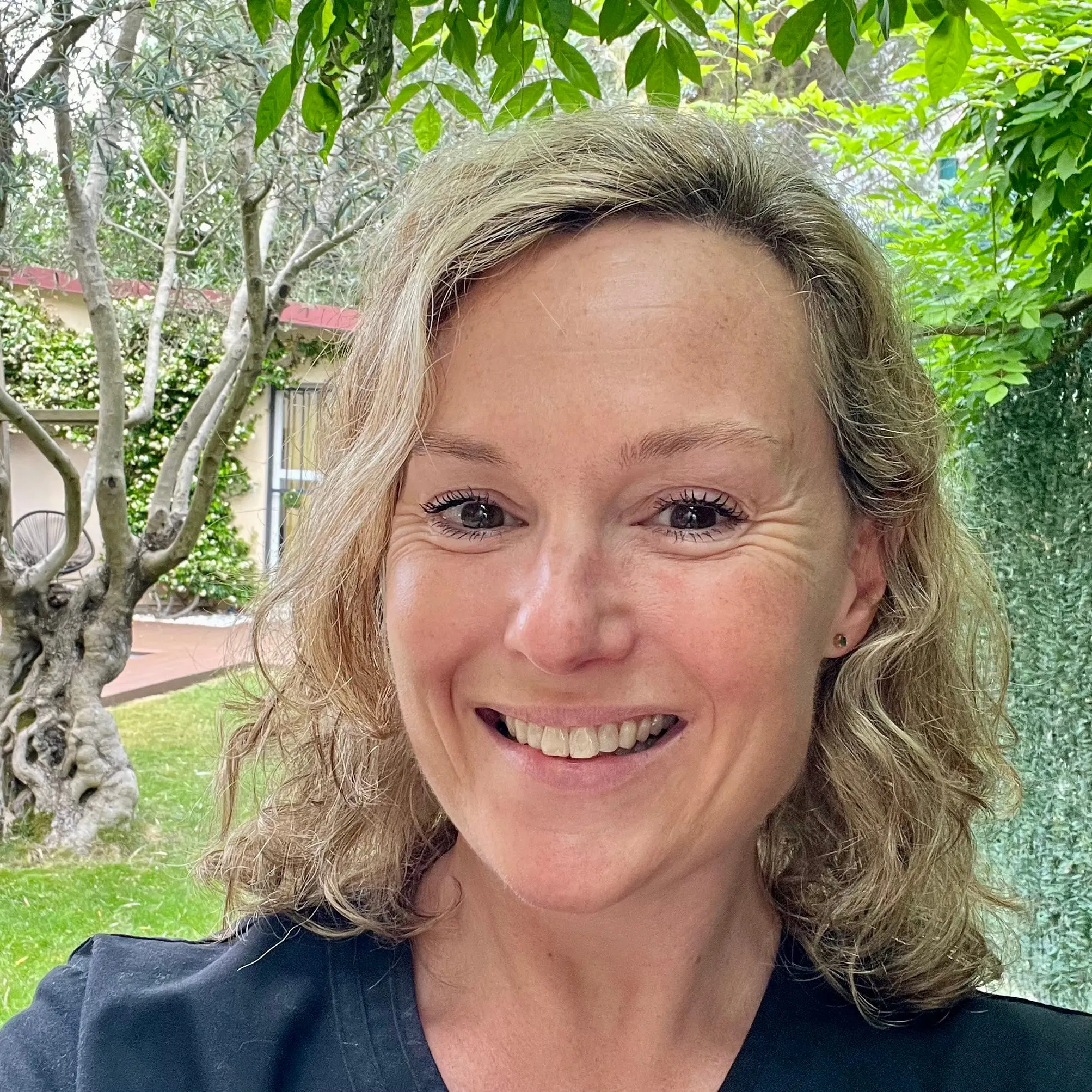 Portrait souriant d’une femme aux cheveux blonds ondulés, en tenue sombre, devant un olivier et un jardin verdoyant.