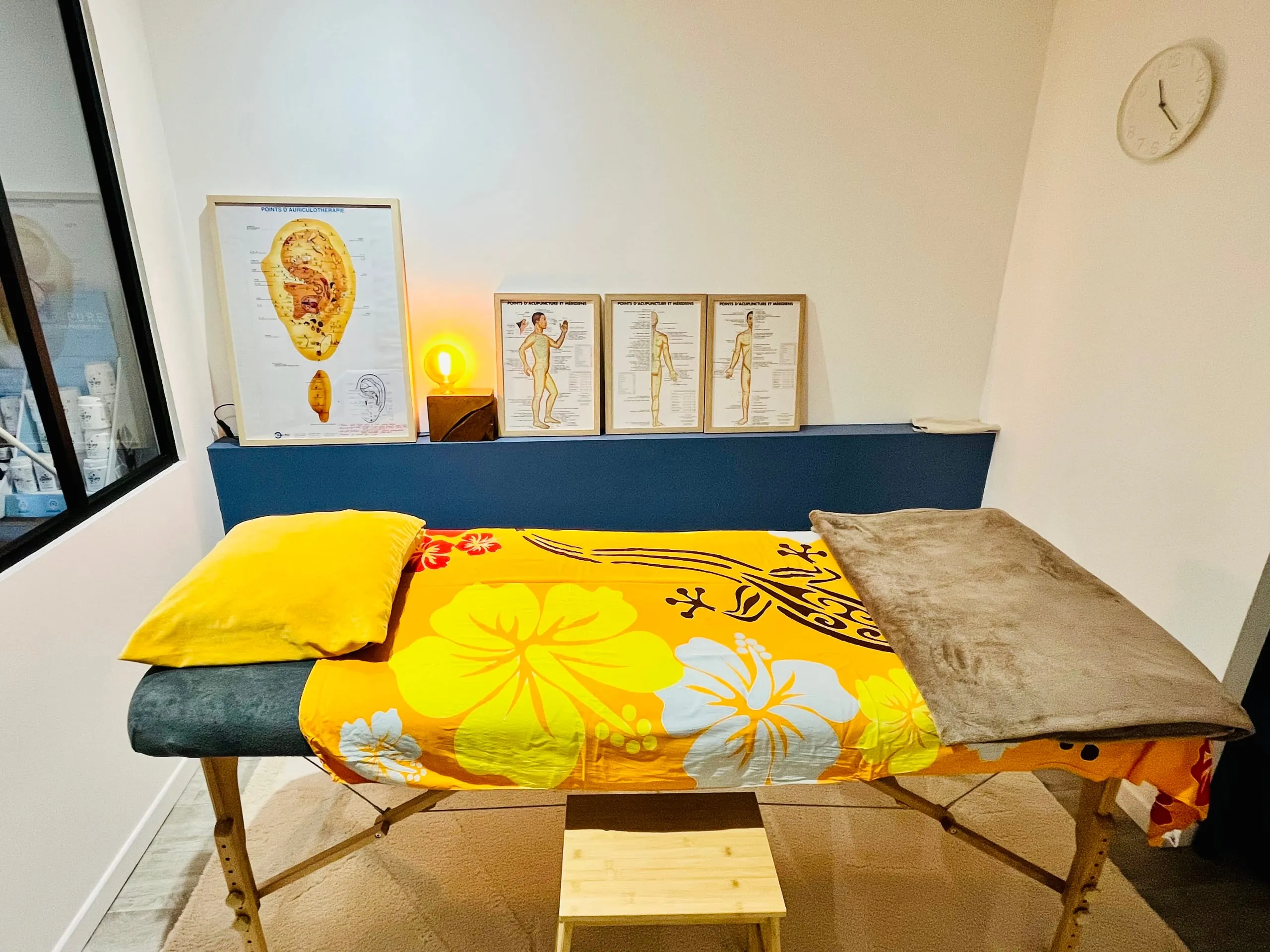 Salle de soin lumineuse avec table de massage recouverte d’un tissu orange à fleurs d’hibiscus, planches d’auriculothérapie au mur et lampe chaude.