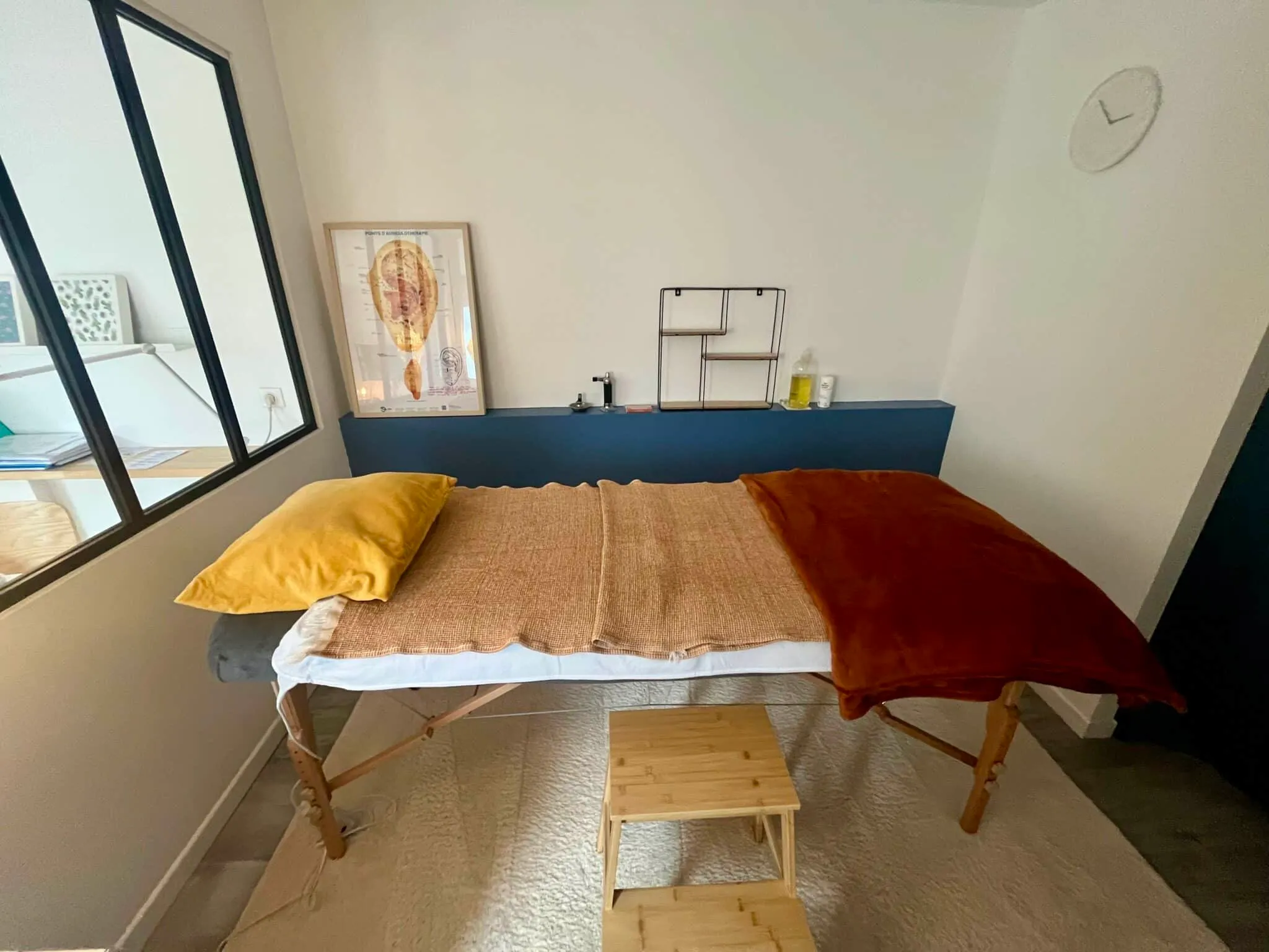Cabinet de soin lumineux avec table de massage, coussins colorés et schéma d’auriculothérapie encadré.