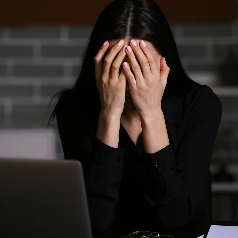 Femme aux cheveux noirs, mains sur le visage, visiblement stressée ou épuisée devant un ordinateur portable, dans un environnement sombre.