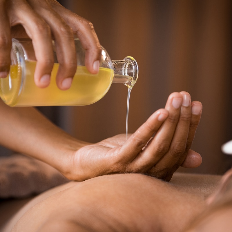 Personne versant de l'huile de massage dorée d'une bouteille en verre dans une main ouverte, préparant un soin spa relaxant.