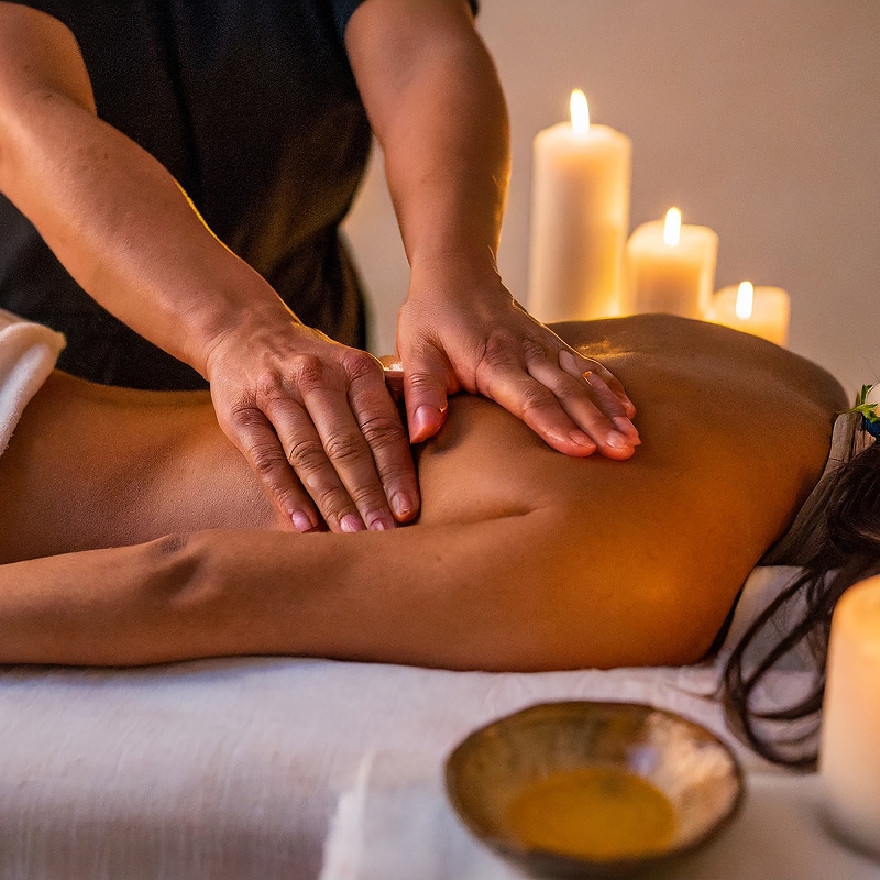 Mains d'un masseur prodiguant un massage du dos apaisant à une personne allongée, ambiance relaxante avec bougies et huile.