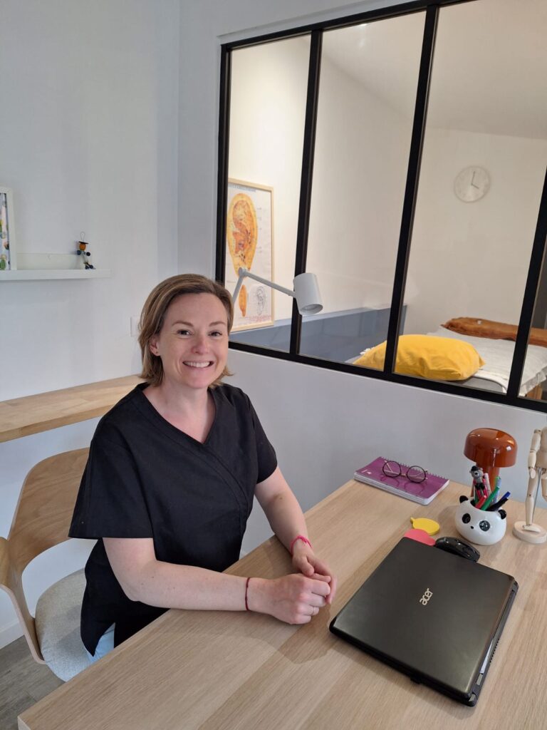 Femme souriante en tenue médicale noire assise à son bureau, avec un ordinateur Acer, une affiche anatomique et une salle de soin derrière.