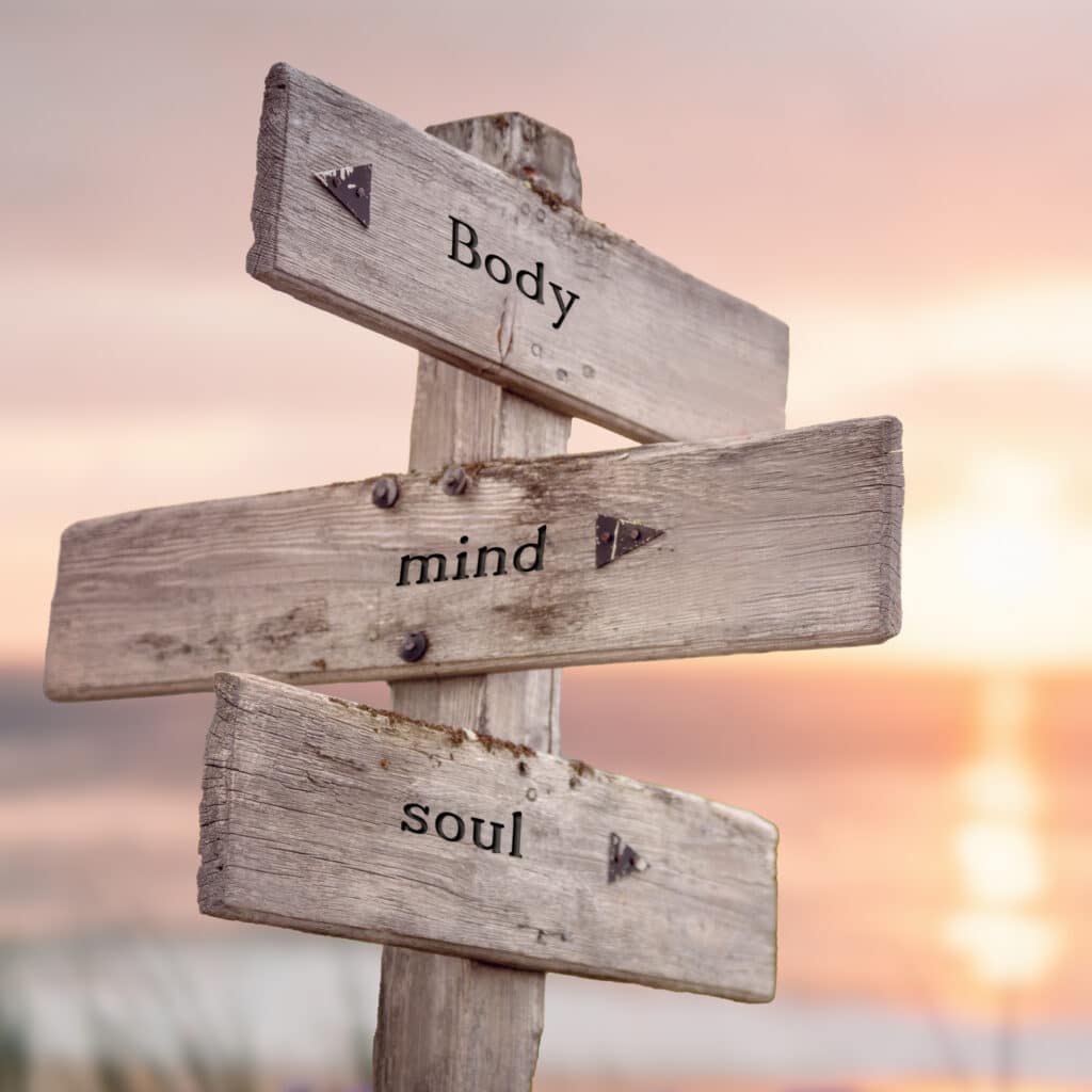 Panneau en bois avec flèches indiquant 'Body', 'mind' et 'soul' sur fond de ciel au lever/coucher de soleil orangé. Symbole de bien-être.