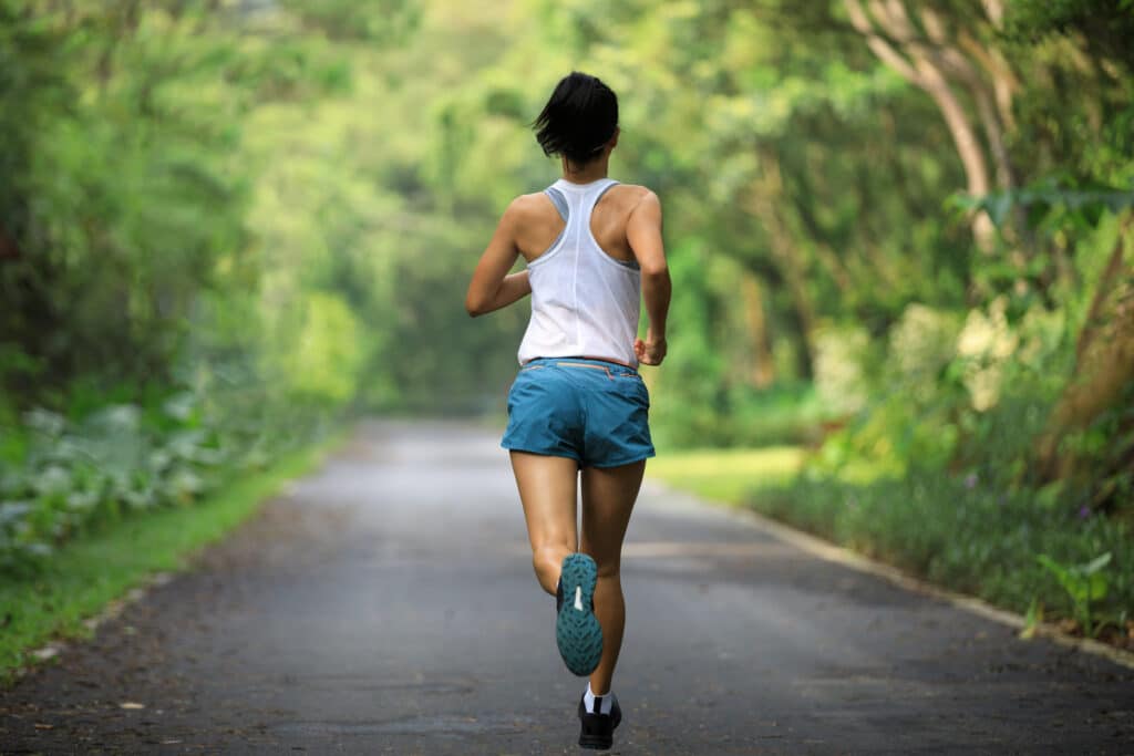 Femme athlétique de dos, en tenue de sport blanche et bleue, courant sur un chemin bordé d'arbres. Course en pleine nature.