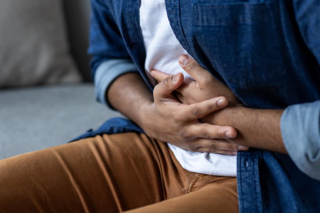 Homme en chemise bleue et pantalon marron, se tenant le ventre des deux mains, signe de douleur abdominale ou indigestion.