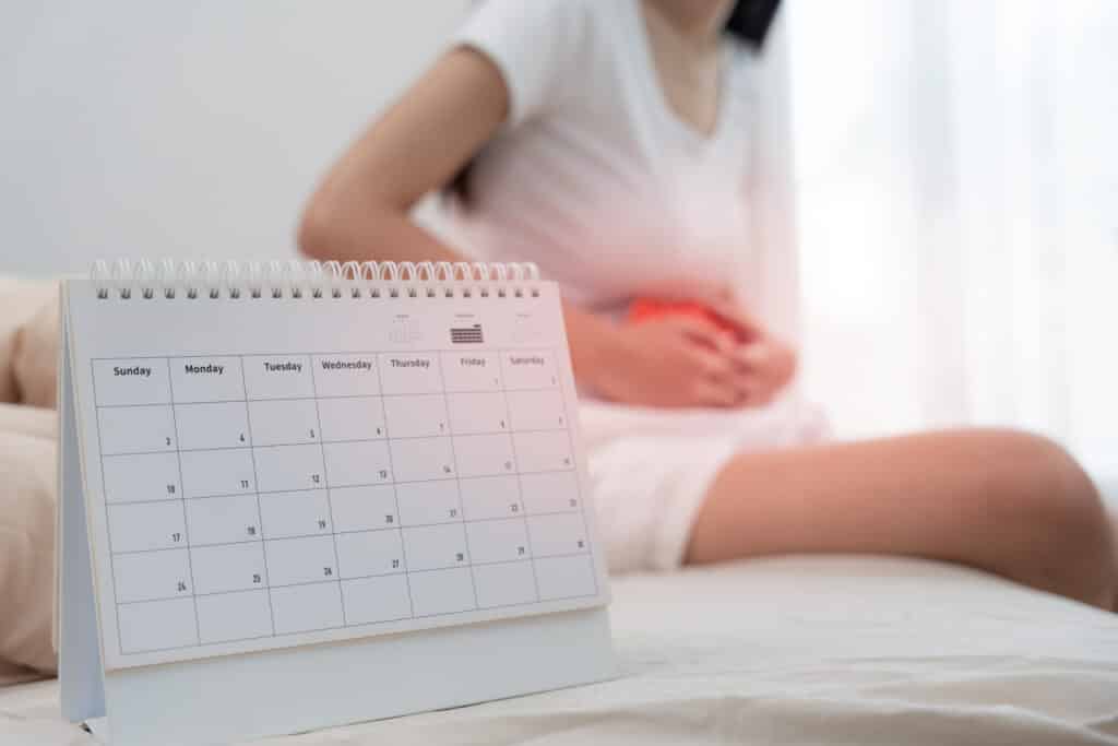 Calendrier de bureau affichant août (3-30). Femme en arrière-plan, assise, tenant son ventre, suggérant des douleurs menstruelles.