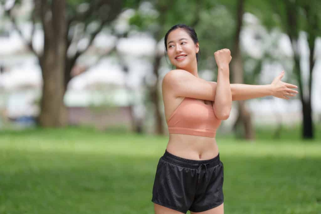 Jeune femme asiatique souriante en tenue de sport orange et noire, étirant son bras dans un parc verdoyant.