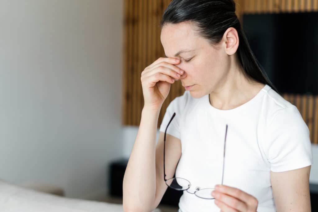 Femme fatiguée se frottant le pont du nez, tenant ses lunettes, indiquant fatigue oculaire ou mal de tête. Cheveux sombres, t-shirt blanc.