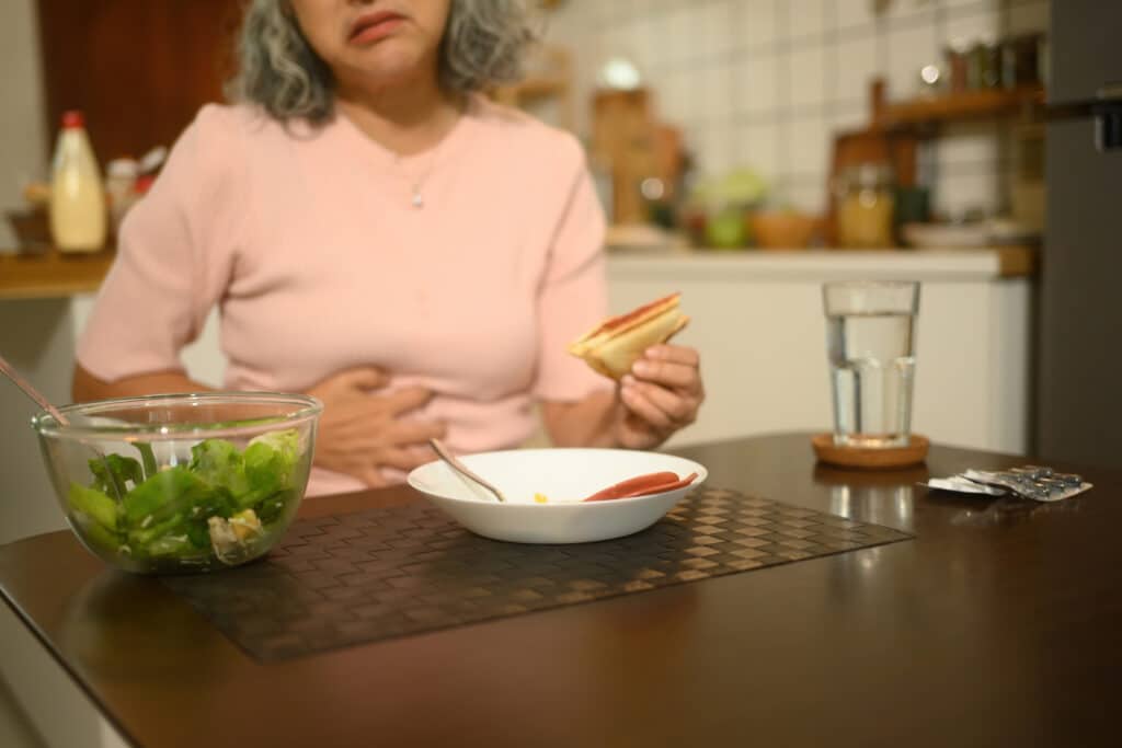 Femme âgée aux cheveux gris souffrant de maux d'estomac, main sur le ventre, tenant un sandwich dans une cuisine. Salade, eau et pilules sur la table.