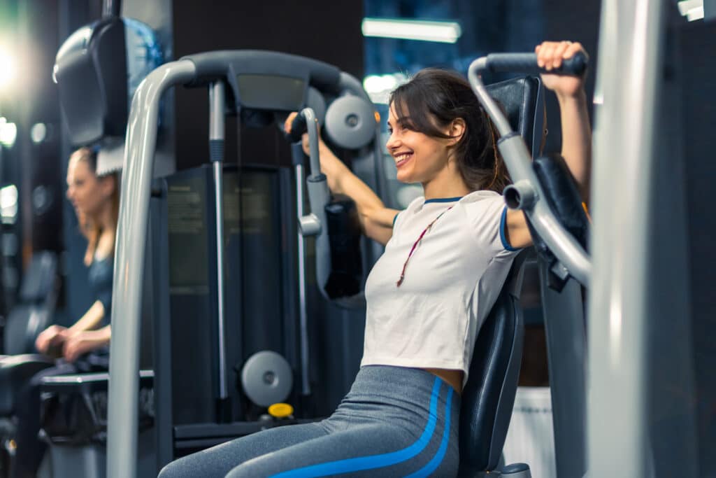 Jeune femme souriante faisant de la musculation sur une machine de presse poitrine en salle de sport.