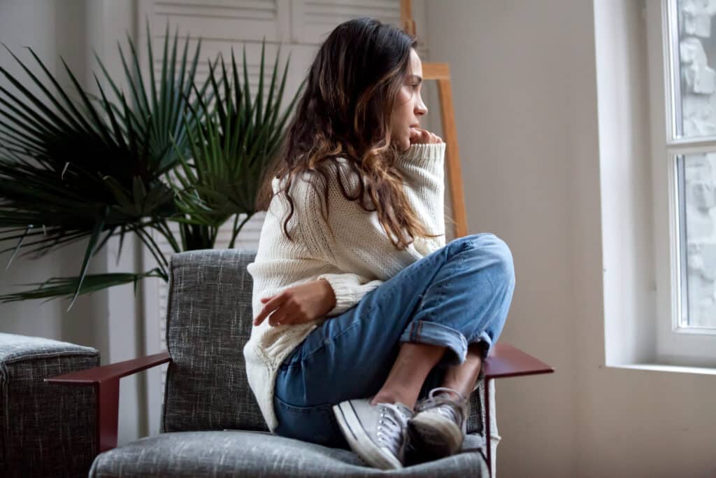 Femme pensive à la fenêtre | Moments de calme et réflexion Jeune femme pensive en pull blanc et jean, assise sur un fauteuil, regardant par la fenêtre. Ambiance calme et introspective.