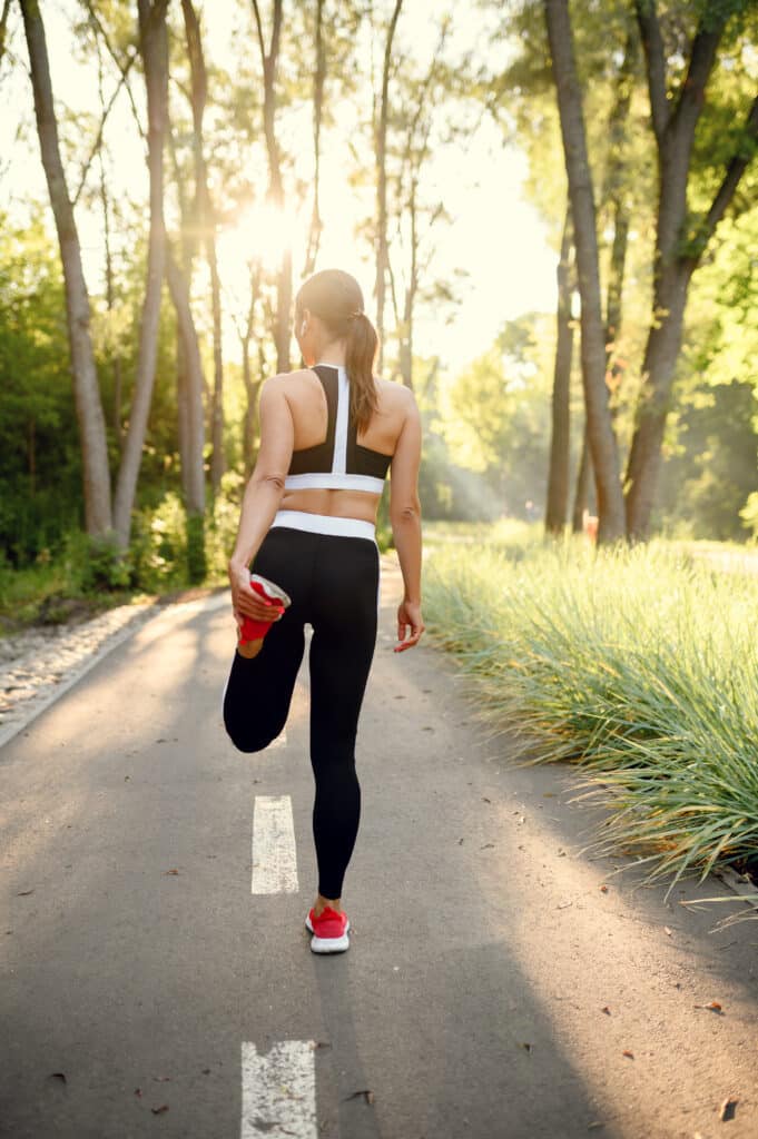 Femme en tenue de sport noire et blanche s'étire la jambe sur un chemin ensoleillé. Arbres et nature verdoyante au coucher du soleil.
