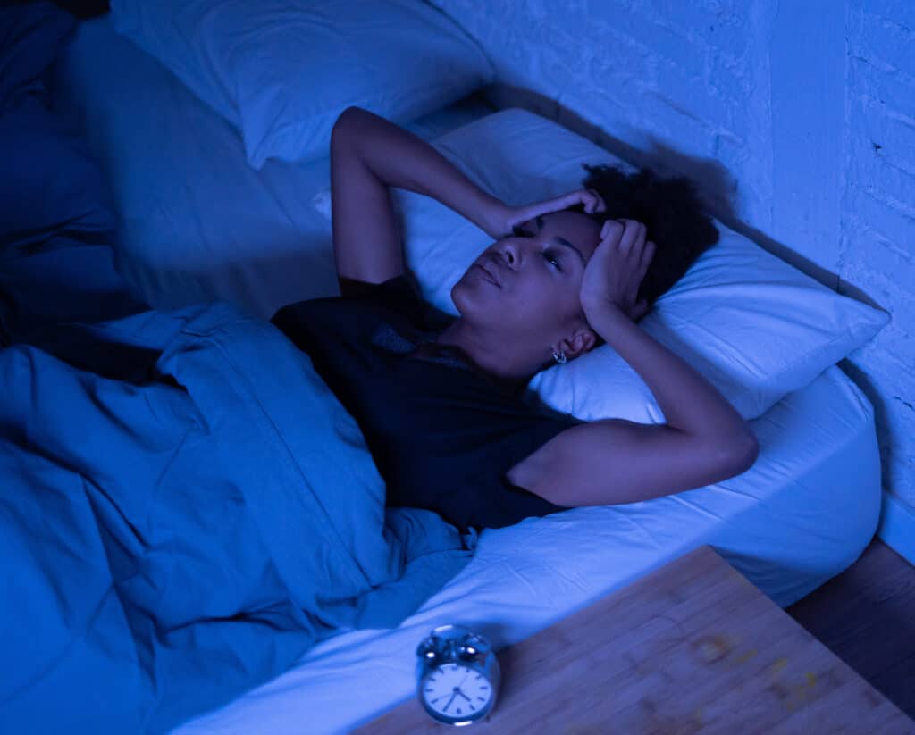 Femme souffrant d'insomnie, allongée dans un lit bleu la nuit, se tenant la tête. Un réveil matinal est sur la table de chevet.