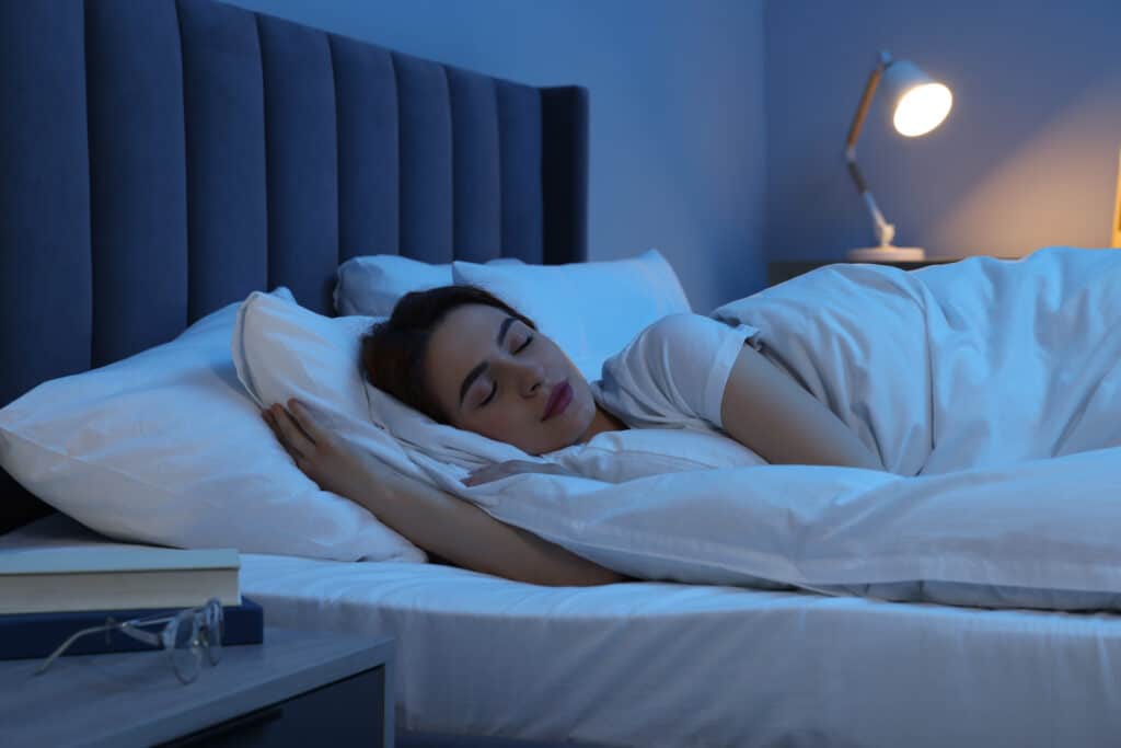 Femme dormant paisiblement sur le côté dans un lit confortable avec tête de lit bleue et draps blancs. Lampe de chevet allumée.
